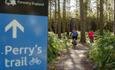 Image shows the Perry's Trail sign at Cannock Chase Forest, with three mountain-bikers zooming off into the distance